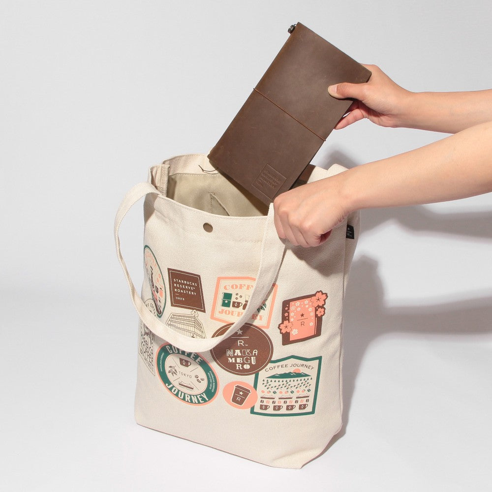 A cream canvas tote bag with colorful travel-style patch prints on the front, shown with a person's hands inserting a brown leather notebook or folder into the open bag against a clean white background.